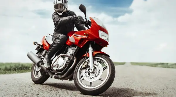 Motociclista con casco e giacca in pelle, in posizione di guida sportiva su una moto rossa su una strada vuota.
