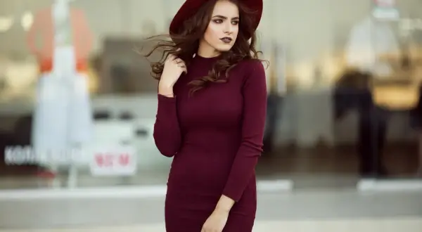 Elegancka bordowa sukienka i kapelusz tworzą stylową stylizację. Kobieta pozuje na tle witryny sklepowej.