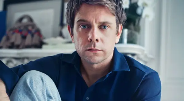 Ritratto di Jonathan Anderson, con occhi azzurri penetranti, indossa una camicia blu e jeans.