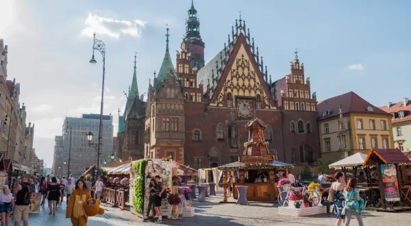 Tętniący życiem wrocławski Rynek, z Ratuszem w tle. Ludzie spacerują wśród kramów, a ktoś w stroju misia dodaje uroku temu miejscu.