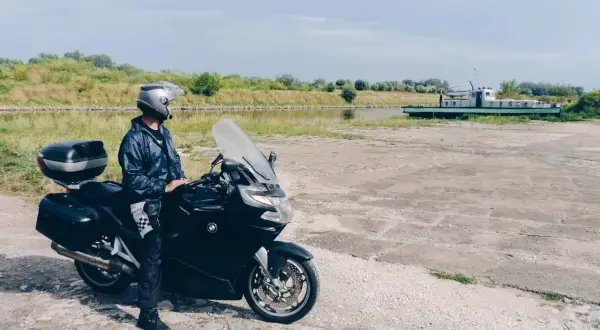 Motocyklista w kasku i kurtce stoi obok czarnego motocykla nad kanałem z łodzią w tle.