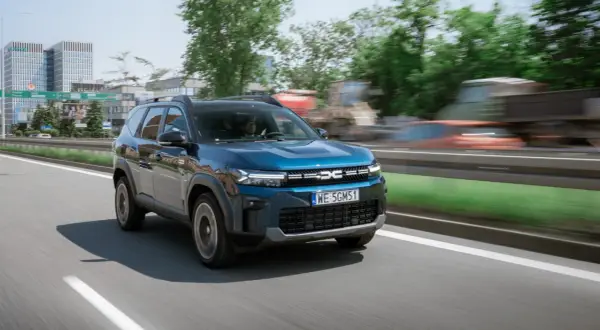 Niebieski Dacia Jogger na drodze, z widocznymi budynkami w tle. Marka samochodu na m.