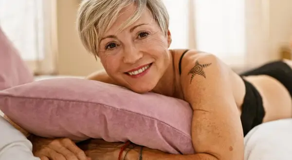 Lächelnde Frau mit kurzem blonden Haar und einem Stern-Tattoo auf der Schulter liegt auf einem Kissen.