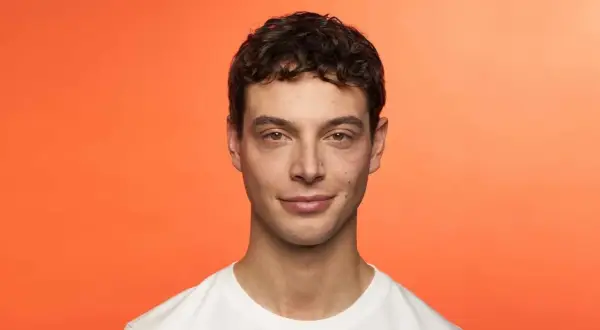 Hombre joven con cabello castaño rizado, luciendo una permanente ondulada. Sonríe levemente ante un fondo naranja vibrante.