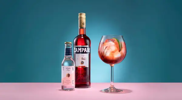 En Campari Spritz med is och lime i ett stort glas. Bredvid står en flaska Campari och en flaska tonic.