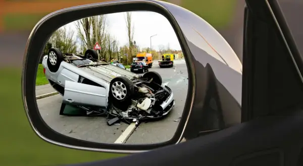 Auto w leasingu leży na dachu po wypadku. Widok w lusterku bocznym pokazuje szkody z OC sprawcy i pomoc drogową na drodze.