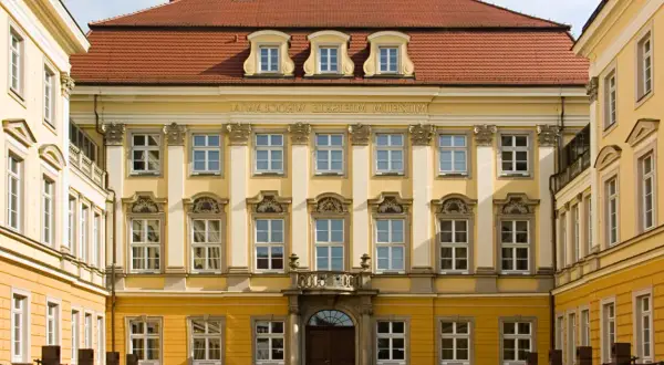 Elegancki budynek z żółtą fasadą i czerwonym dachem, ozdobiony licznymi oknami. To muzeum sztuki medalierskiej we Wrocławiu.