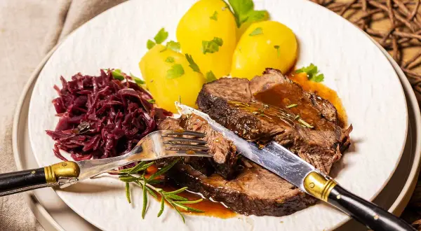Zarter Rinderbraten im Ofen, serviert mit Salzkartoffeln und Rotkohl. Ein Messer und eine Gabel sind bereit, das saftige Fleisch anzuschneiden.