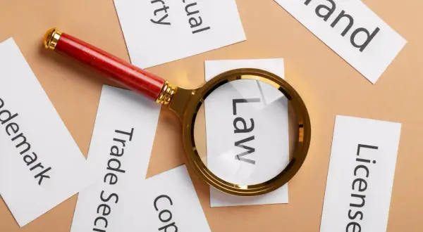 Lupa powiększa słowo "Law" wśród kart z napisami: "Brand", "License", "Copyright", "Trade Secret", "Trademark", "Intellectual Property". To symboliczne przedstawienie analizy prawnej wartości niematerialnych i prawnych.