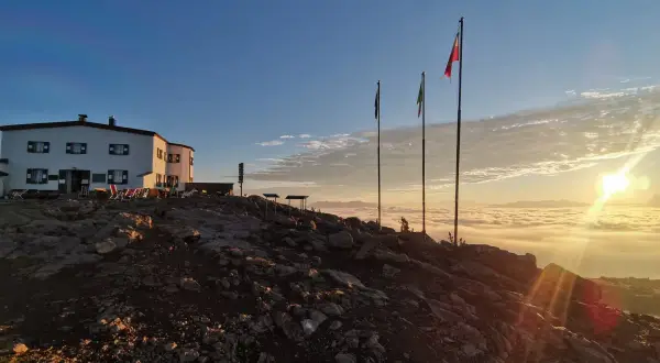 Sonnenaufgang über einer Wolkendecke, mit Hütte und Fahnen. Ein perfekter Moment auf der Sarntaler Hufeisentour.