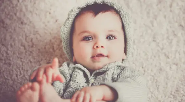 Bebé con ojos grandes y azules, vistiendo un gorro y suéter tejidos. Podría llamarse Gabriel, un nombre con g.