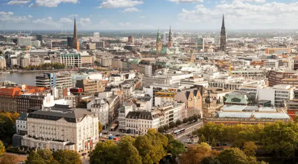 Blick von einer **Aussichtsplattform Hamburg** über die Stadt mit ihren Kirchtürmen, dem Hafen und grünen Bäumen im Vordergrund.