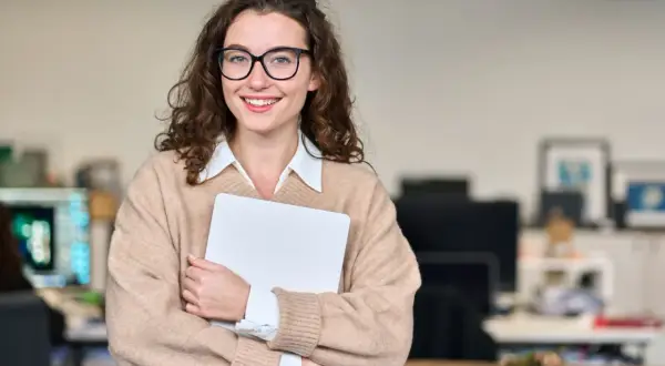 Uśmiechnięta kobieta w okularach trzyma laptopa. Jej strój i pewność siebie sugerują, że doskonale odnajduje się w pracy związanej z obsługą klienta, kod zawodu.