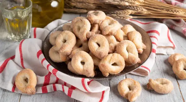 Piatto pieno di taralli dolci al vino bianco, cosparsi di zucchero. Accanto, un bicchiere di vino bianco e una bottiglia d'olio.