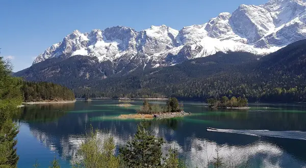 Idyllische Bergkulisse mit schneebedeckten Gipfeln, einem türkisfarbenen See und kleinen Inseln. Ein Boot gleitet über das Wasser, ideal für den Kramerplateauweg Rundweg.