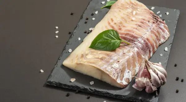 Surowy filet z dorsza czarnego, doprawiony solą i pieprzem, z czosnkiem i listkami bazylii na czarnej desce.