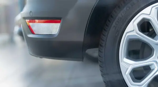 Vue rapprochée du pare-chocs arrière d'une voiture blanche, montrant un catadioptre rouge et blanc, un capteur de stationnement et une roue avec jante argentée. Le catadioptre voiture obligatoire assure la sécurité.