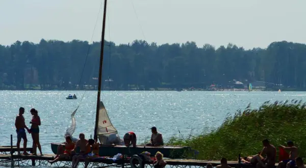 Ludzie wypoczywają na pomoście nad jeziorem, w tle las i żaglówki. Pojezierze Łęczyńsko-Włodawskie tętni życiem.