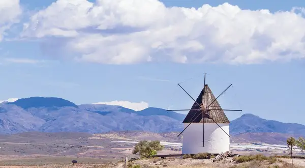Molino blanco con aspas de madera bajo un cielo azul con nubes. Paisaje árido con montañas al fondo, como si Fernan Perez hubiera pintado este cuadro.