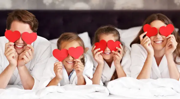 Familia feliz celebra San Valentín para niños, cubriendo sus ojos con corazones rojos en la cama.