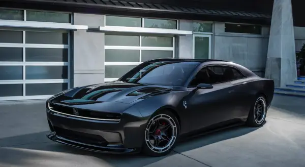 Nowoczesny, ciemnoszary Dodge Daytona z charakterystycznym pasem LED z przodu, zaparkowany przed nowoczesnym budynkiem z garażem.
