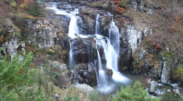 Il sentiero delle cascate serpeggia tra rocce e foliage autunnale, con l'acqua che scende impetuosa.