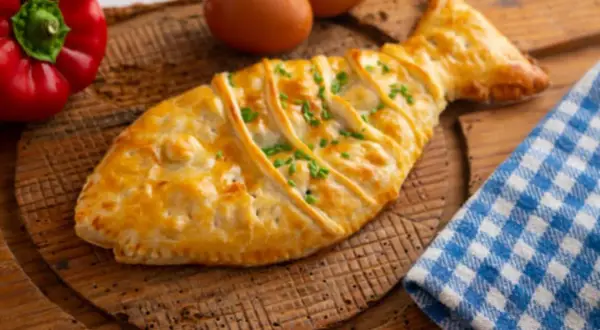 Empanada de bacalao con forma de pez, dorada y adornada con cebollino. Acompañada de pimiento rojo y huevos.