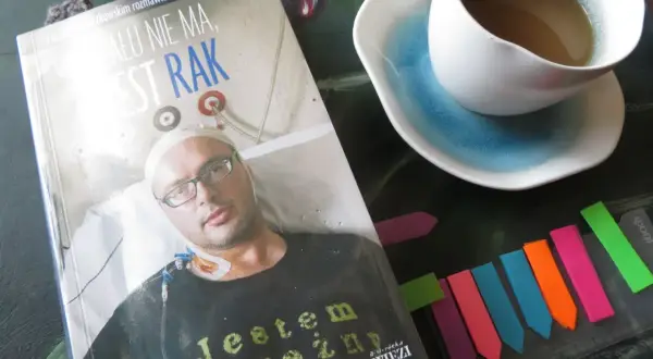 Książka "Żalu nie ma, jest rak" z wizerunkiem pacjenta, obok filiżanka kawy i kolorowe zakładki.