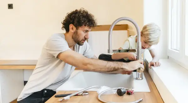 Padre e figlio installano un nuovo rubinetto in cucina, un'operazione che richiede attenzione per spostare attacchi cucina senza rompere pavimento.