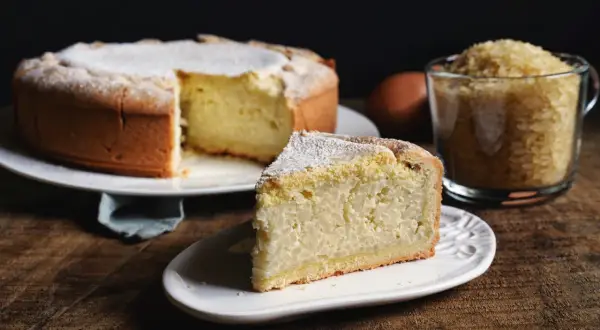 Una fetta di torta di riso, la ricetta originale, cosparsa di zucchero a velo, accanto alla torta intera e a una tazza di riso.