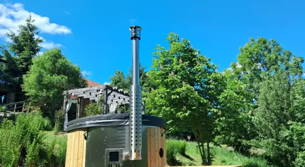 Drewniane jacuzzi w ziemi z metalowym kominem, otoczone zielenią i błękitnym niebem. Idealne miejsce na relaks.