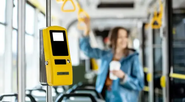 Żółty biletomat w autobusie, obok siedzeń. Kobieta w tle trzyma kubek. Pomyśl o zniżkach i ulgach na przejazdy.
