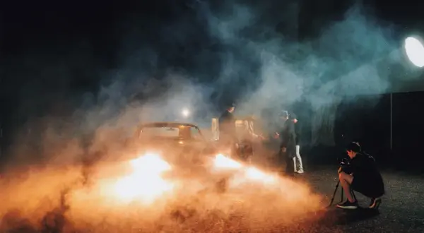 A film crew uses artificial light examples, including a bright spotlight and car headlights, to create a smoky, dramatic scene with men in hats gathered around a vintage car.