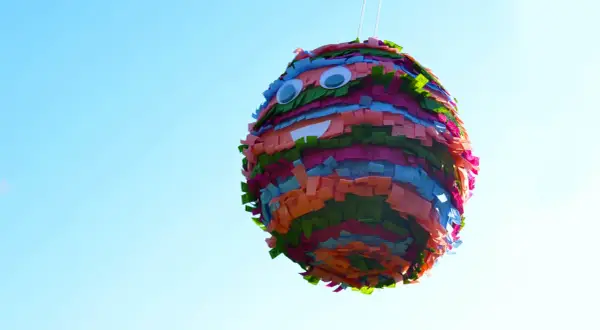 Piñata casera colorida con ojos saltones y boca sonriente, colgada contra un cielo azul claro.