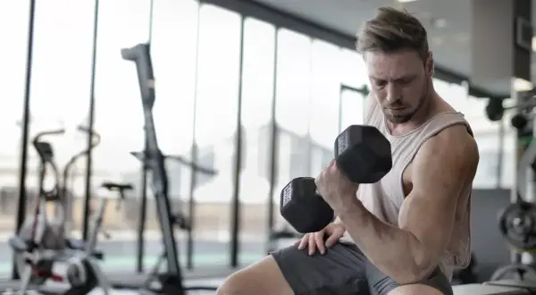 Mężczyzna ćwiczy biceps z hantlem, pokazując mięśnie synergistyczne przykłady. W tle siłownia.