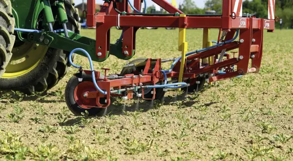 Un tracteur vert et un pulvérisateur rouge travaillent dans un champ de jeunes pousses. La machine applique un traitement ciblé, peut-être pour une liste de graminées à éradiquer.