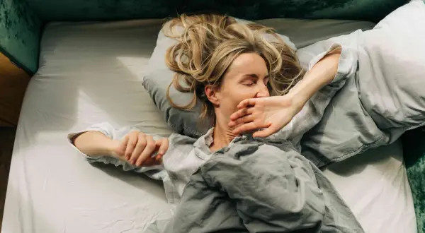 Woman in bed, covering her mouth, looking exhausted. Could this be narcolepsy vs hypersomnia?
