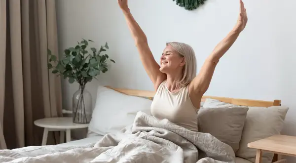 A woman stretches her arms up, smiling, in bed. A perfect way to learn how to wake yourself up feeling refreshed and ready for the day.