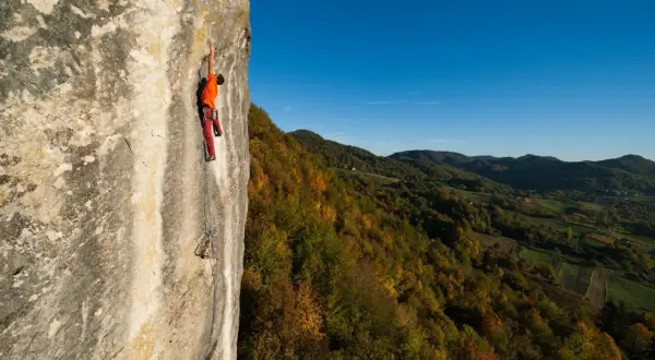 Ein Kletterer in orangefarbenem Shirt und roten Hosen erklimmt eine Felswand in Kroatien, während sich im Hintergrund ein herbstliches Tal erstreckt.