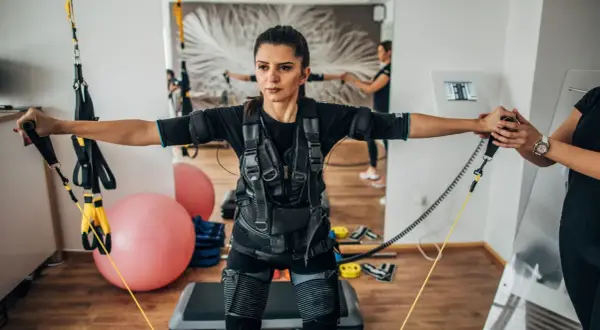 Kobieta w kamizelce do treningu EMS wykonuje ćwiczenia z taśmami. W tle widać drugą kobietę i sprzęt do ćwiczeń.