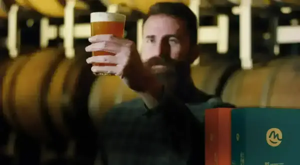 Uomo con barba esamina un bicchiere di birra dorata, con botti di legno sullo sfondo. Scatole di birre lager famose in primo piano.