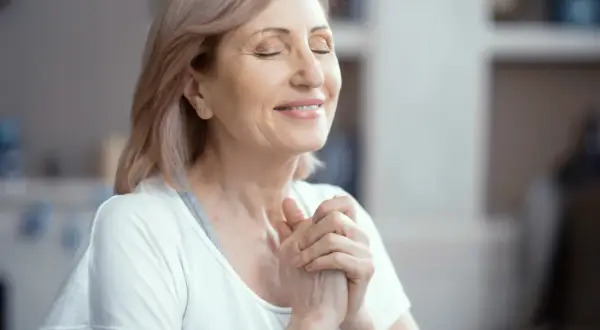 Uśmiechnięta kobieta z zamkniętymi oczami, dłonie złożone na piersi, praktykuje techniki mindfulness.