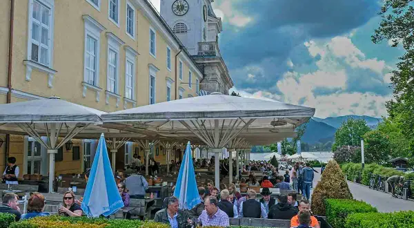 Menschen genießen die Tegernsee Feste unter Sonnenschirmen. Ein Gebäude mit Turmuhr und bayerische Fahnen im Hintergrund.