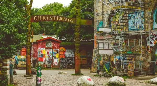 Drewniany totem i tablica z napisem "CHRISTIANIA" wita w tej wolnej dzielnicy. Obok czerwony budynek z plakatami i rowery.