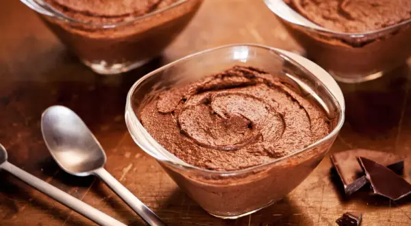 Trois coupelles de mousse au chocolat sans oeuf, onctueuse et gourmande, avec des morceaux de chocolat noir et des cuillères sur fond de bois.