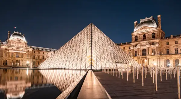 Das Louvre-Museum in Paris bei Nacht, mit der beleuchteten Glaspyramide im Vordergrund und dem historischen Gebäude im Hintergrund.