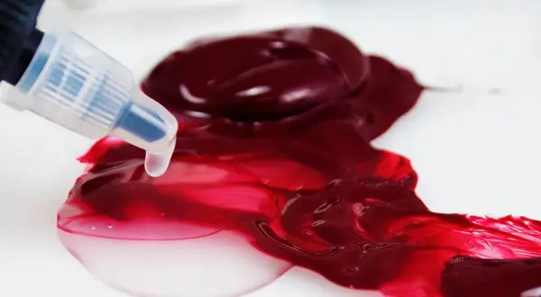Un beccuccio trasparente rilascia un liquido chiaro su una macchia di vernice acrilica color bordeaux, creando sfumature vivaci.