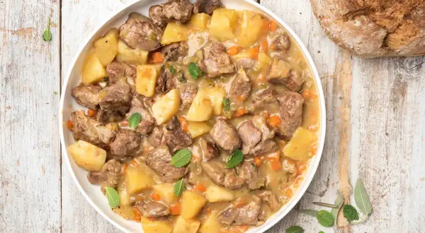 Un saporito agnello in umido con patate, carote e erbe aromatiche, servito in un piatto bianco su un tavolo di legno rustico.