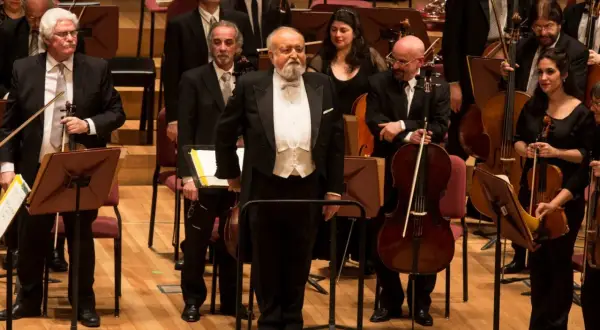 Orkiestra przygotowuje się do wykonania Dies Irae. Dyrygent w białej kamizelce i fraku stoi na podwyższeniu, otoczony muzykami z instrumentami smyczkowymi.