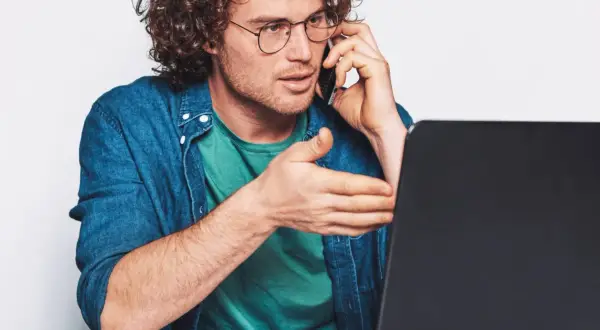 Mężczyzna w okularach rozmawia przez telefon, gestykulując w stronę laptopa. Szuka uczciwych firm oddłużeniowych, by rozwiązać swoje problemy finansowe.